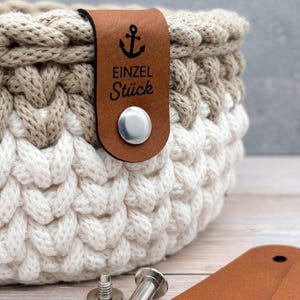 May include: A round, two-toned crochet basket featuring a thick, braided knit texture in beige and white. A brown leather tag is attached to the rim with a silver metal rivet, displaying an anchor icon and the text EINZEL St&uuml;ck.