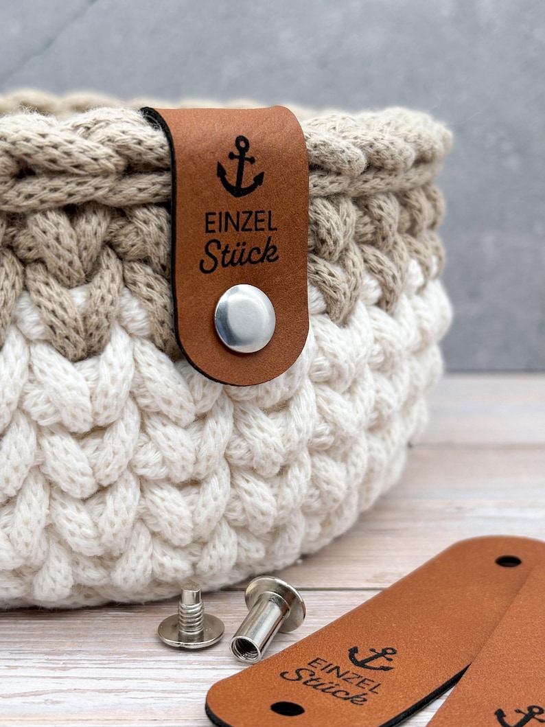 May include: A round, two-toned crochet basket featuring a thick, braided knit texture in beige and white. A brown leather tag is attached to the rim with a silver metal rivet, displaying an anchor icon and the text EINZEL St&uuml;ck.