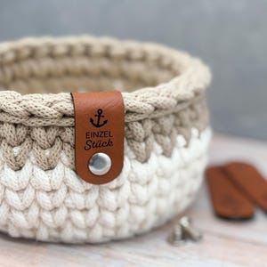 May include: A round, hand-crocheted basket featuring a two-tone design with a white base and a beige upper section. A brown faux leather tag is attached to the rim with a silver metal snap, displaying an anchor icon and the text EINZEL St&uuml;ck.