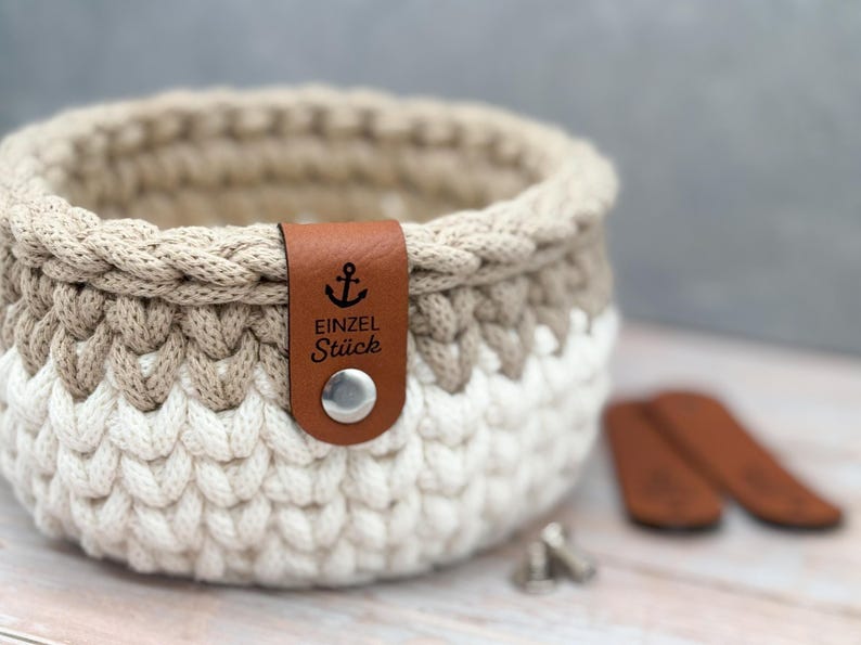 May include: A round, hand-crocheted basket featuring a two-tone design with a white base and a beige upper section. A brown faux leather tag is attached to the rim with a silver metal snap, displaying an anchor icon and the text EINZEL St&uuml;ck.