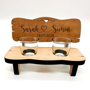 May include: Wooden wedding shot glass holder with two shot glasses. The holder is shaped like a bench and has a sign that says "Sarah & Simon 14.09.2024".