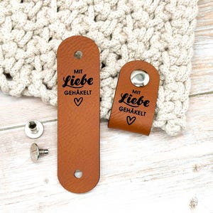 Könnte beinhalten: Zwei hellbraune Lederetiketten mit abgerundeten Enden, bedruckt mit dem schwarzen Text "MIT LIEBE GEHÄKELT" und einem Herzsymbol. Ein Etikett ist flach mit zwei Löchern, das andere ist gefaltet und mit einem silbernen Metalldruckknopf versehen. Zwei Metallnieten liegen bei.