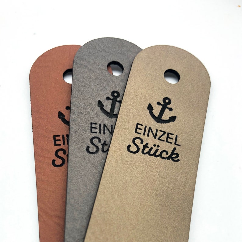 May include: Three rectangular tags with rounded tops and a circular hole at the top of each. The tags are brown, dark grey, and light tan. Each tag features a black printed anchor icon above the German text EINZEL ST&Uuml;CK in a sans-serif font.