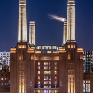 Può includere: Un edificio in mattoni con quattro alti camini illuminati di notte. L'edificio ha molte finestre ed è illuminato da luci gialle calde. L'edificio è un ex centrale elettrica che è stata convertita in una galleria d'arte moderna.