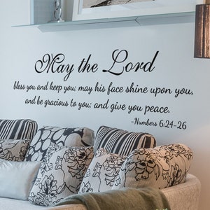 Puede incluir: Una calcomanía de pared con una fuente de escritura negra en cursiva que dice "Que el Señor te bendiga y te guarde; que su rostro brille sobre ti, y te sea propicio; y te dé paz. -Números 6:24-26". La calcomanía está en una pared blanca sobre un sofá beige con almohadas de patrones negros y blancos.
