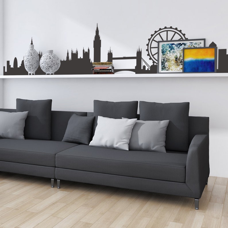 May include: A gray sectional sofa with white and gray pillows in front of a white wall with a black silhouette of the London skyline, including Big Ben, the London Eye, and Tower Bridge. Two decorative vases are on a white shelf below the silhouette.
