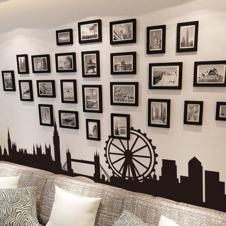 May include: A white wall with a black and white photo collage of various landmarks and cityscapes. The photos are in black frames and are arranged in a random pattern. The collage is above a black silhouette of the London skyline, including the London Eye and Big Ben.