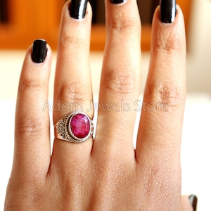 May include: A silver ring with a pink oval gemstone set in a decorative silver band. The ring is on a person's finger.