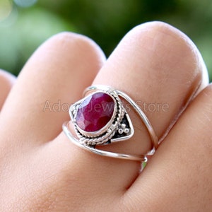 May include: A silver ring with a red ruby gemstone set in a delicate, ornate design. The ring features a twisted band and a small, triangular accent.