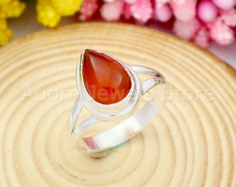 May include: A silver ring with a teardrop-shaped carnelian gemstone. The ring is set on a wooden surface.