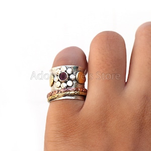 May include: A silver-toned spinner ring with a central cluster of small, round silver accents and a single, round, deep red gemstone. The ring features gold and copper bands, and is worn on a finger.