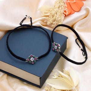 Op de afbeelding: Twee zwarte koordarmbanden met vierkante, juweelkleurige middelen. Elk middelpunt heeft een vierkante witte edelsteen omringd door kleinere rode en groene edelstenen. De armbanden worden gepresenteerd op een blauw boek.