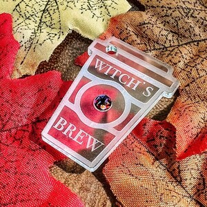 May include: A clear acrylic keychain shaped like a coffee cup with the text "Witch's Brew" and a red rhinestone in the center. The keychain is on a background of fall leaves.