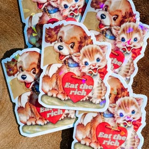 Puede incluir: Cinco pegatinas con un perro y un gato de dibujos animados que sostienen un corazón rojo con el texto "Eat the rich". Las pegatinas son holográficas y tienen una estética vintage.