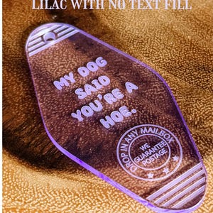 May include: A translucent lilac motel-style keychain with a rounded diamond shape. The text reads: MY DOG SAID YOU'RE A HOE. Below this, a circular emblem features the text: DROP IN ANY MAILBOX WE GUARANTEE POSTAGE. It has a hole at the top.