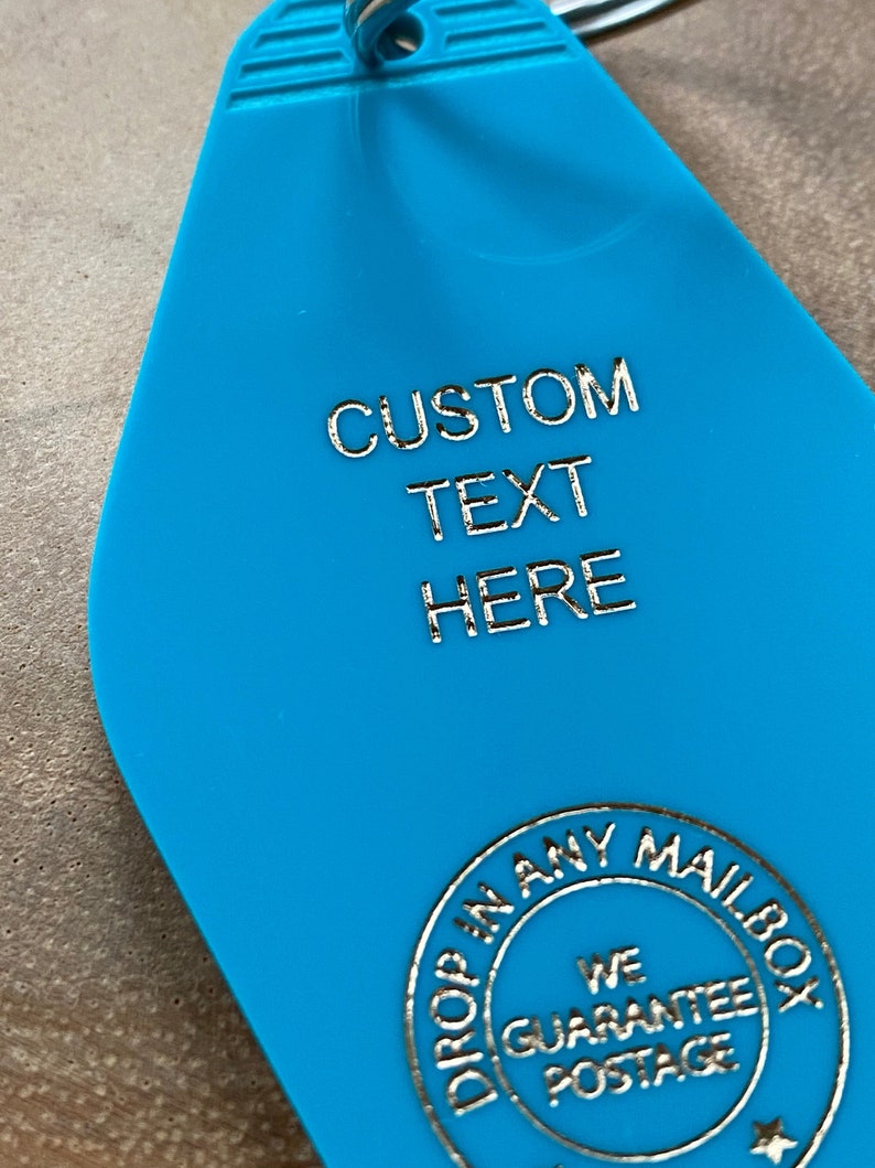 May include: A bright blue motel-style keychain with a teardrop shape and a silver metal ring. The surface features the embossed silver text CUSTOM TEXT HERE and a circular emblem that reads DROP IN ANY MAILBOX WE GUARANTEE POSTAGE.