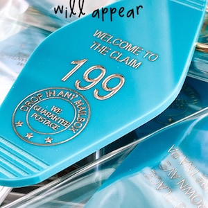 May include: A teal motel-style keychain with gold-colored text reading "WELCOME TO THE CLAIM 199" and a circular emblem stating "DROP IN ANY MAILBOX WE GUARANTEE POSTAGE" with three stars. The text "this is how room numbers will appear" is visible.