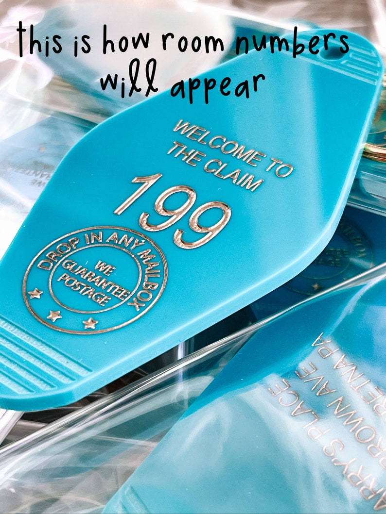 May include: A teal motel-style keychain with gold-colored text reading "WELCOME TO THE CLAIM 199" and a circular emblem stating "DROP IN ANY MAILBOX WE GUARANTEE POSTAGE" with three stars. The text "this is how room numbers will appear" is visible.
