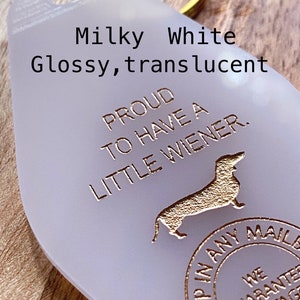 May include: A milky white, translucent, teardrop-shaped motel-style keychain with a gold-colored metal ring. The keychain features the text "PROUD TO HAVE A LITTLE WIENER," a silhouette of a dachshund, and a circular "DROP IN ANY MAILBOX" stamp.