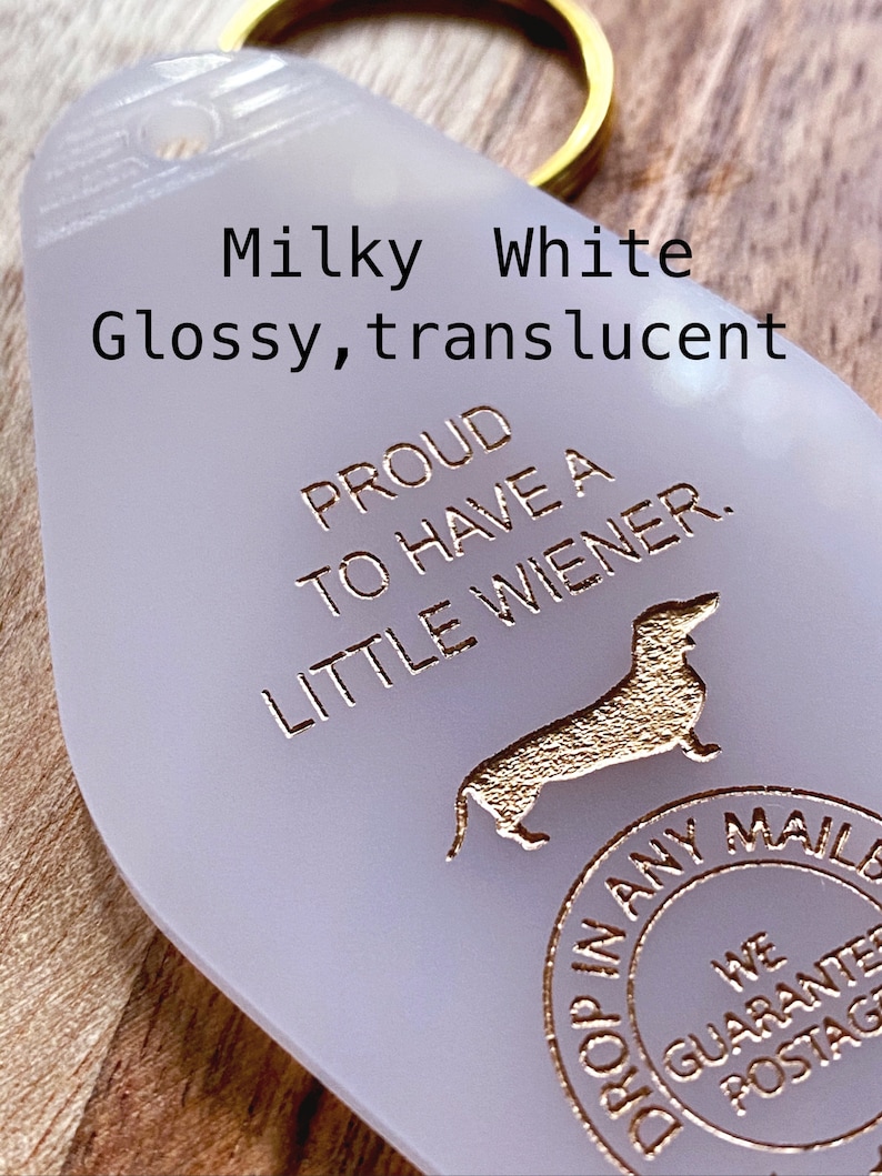 May include: A milky white, translucent, teardrop-shaped motel-style keychain with a gold-colored metal ring. The keychain features the text "PROUD TO HAVE A LITTLE WIENER," a silhouette of a dachshund, and a circular "DROP IN ANY MAILBOX" stamp.