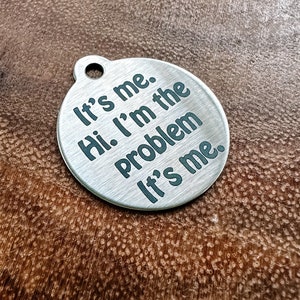 以下が含まれることがあります： 暗い緑色の文字で「It's me. Hi. I'm the problem. It's me.」と刻まれた、丸い銀色の金属タグです。