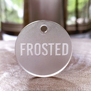 May include: A round, frosted glass tag with a hole at the top and the word "FROSTED" etched in white.