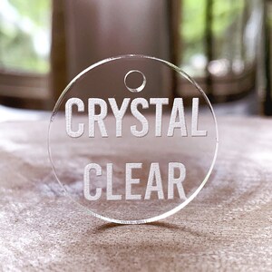 May include: A round, clear acrylic tag with a hole at the top. The tag has the words "CRYSTAL CLEAR" etched in white.
