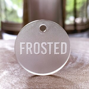 May include: A frosted white round tag with a hole at the top and the word "FROSTED" engraved in white.