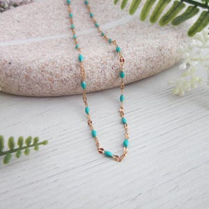 A turquoise-colored necklace with gold-colored beads is displayed on a white surface, surrounded by white flowers and green leaves.