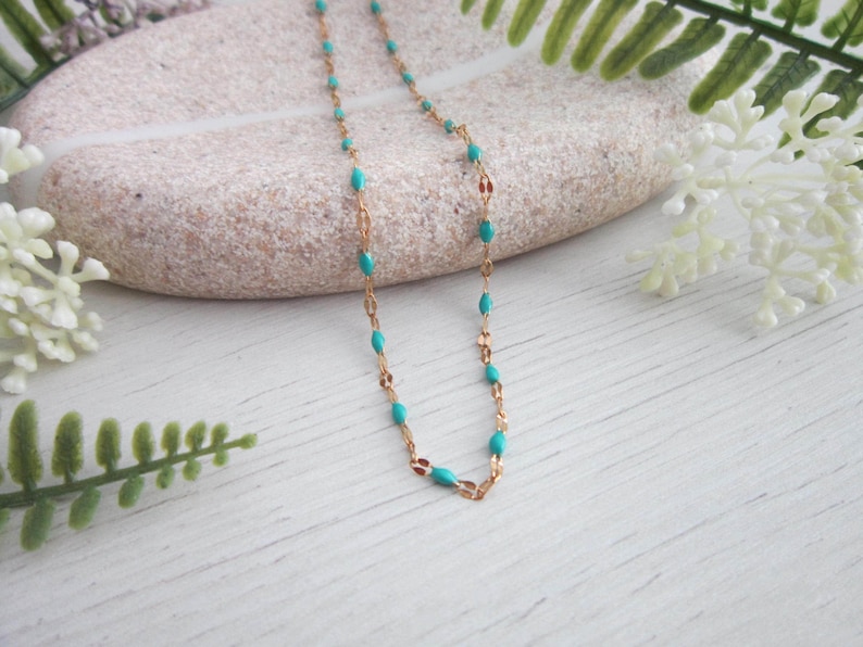 A turquoise-colored necklace with gold-colored beads is displayed on a white surface, surrounded by white flowers and green leaves.