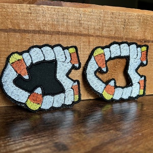 May include: Two embroidered patches shaped like a set of teeth with candy corn in between each tooth. The patches are white, yellow, orange, and black.