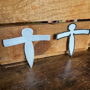 May include: Two white felt patches shaped like stick figures with outstretched arms. The patches are on a wooden surface.