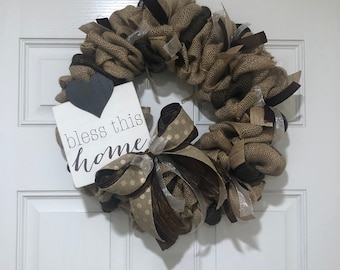 May include: A burlap wreath with a brown and gold ribbon and a sign that says "bless this home".