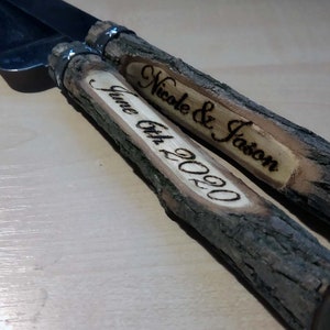 May include: A set of two wooden cake cutting knives with a silver blade. The handles are made of natural wood with the bark still on. The handles are engraved with the text "Nicole & Jason" and "June 6th, 2020".