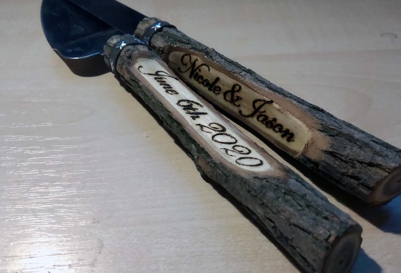 May include: A set of two wooden cake cutting knives with a silver blade. The handles are made of natural wood with the bark still on. The handles are engraved with the text "Nicole & Jason" and "June 6th, 2020".