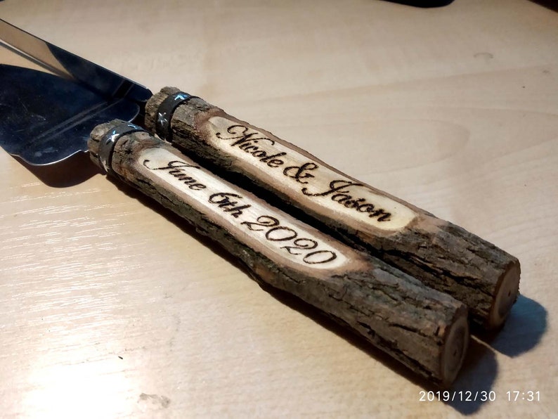 May include: A set of two wooden cake cutting knives with silver bands. The handles are made of natural tree branches and are engraved with the names "Nicole & Jason" and "June 6th 2020".
