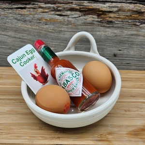 May include: A white ceramic egg cooker with two brown eggs and a bottle of red hot sauce. The hot sauce bottle has a green label with the text "Cajun Egg Cooker" and "Tabasco Brand Pepper Sauce".