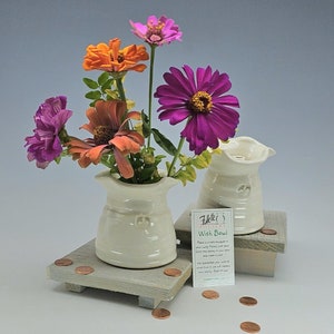May include: Two off-white ceramic wish bowls with a fleur-de-lis design, each holding a bouquet of colorful zinnias. The flowers are in shades of pink, orange, and purple. The bowls sit on a small wooden platform with pennies scattered around.
