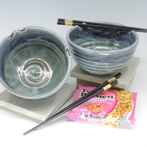 May include: Two handmade ceramic ramen bowls with a blue and green glaze, accompanied by black chopsticks with gold accents. A package of Top Ramen shrimp flavor is also present.