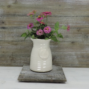 May include: A white ceramic vase with a textured surface and a raised design on the front. The vase is filled with pink flowers.