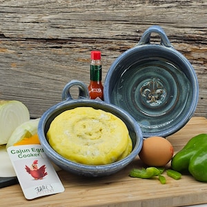 May include: Two blue ceramic Cajun egg cookers with a fleur-de-lis design. One cooker has a yellow egg inside. The cookers are on a wooden surface with a green pepper, a red pepper, and a white onion.