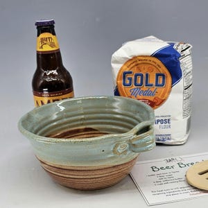 May include: A handmade ceramic bowl with a light blue and brown glaze, a bottle of Abita beer, and a bag of Gold Medal all-purpose flour. A recipe card for beer bread is also visible.