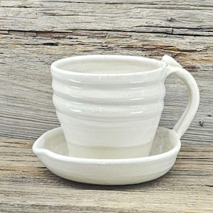 May include: A handmade, off-white ceramic mug with a matching saucer. The mug has a ribbed design and a curved handle. The saucer is attached to the mug. The set is placed on a rustic wooden surface.