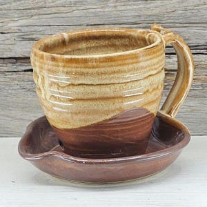 May include: A handmade ceramic mug and saucer set. The mug features a textured, layered design with a light brown and dark brown color scheme. The saucer is a matching dark brown. The mug has a curved handle.