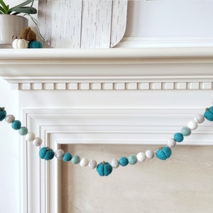 May include: A teal and white felt ball garland with pumpkin shapes hanging over a white fireplace mantel.