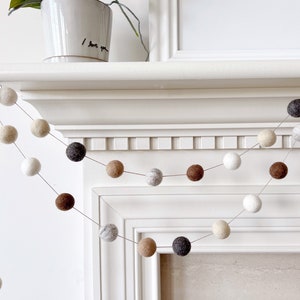May include: A garland of felt balls in shades of brown, beige, white, and gray hangs over a white fireplace mantel. The garland is strung on a clear thread and is approximately 3 feet long.