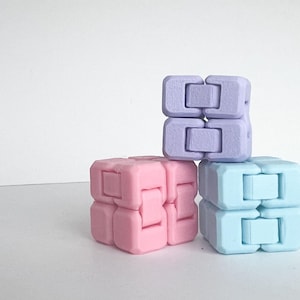 May include: Three colorful fidget cubes in pastel shades of pink, lavender, and light blue. Each cube is composed of two connected blocks. The cubes are stacked on a white surface.