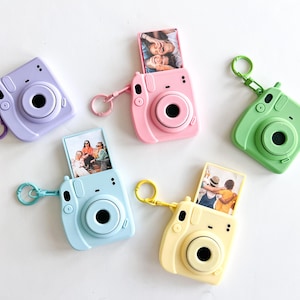 May include: A collection of five miniature camera-shaped keychains in pastel colors: lavender, pink, light blue, green, and yellow. Each keychain holds a small photo and has a matching colored ring.