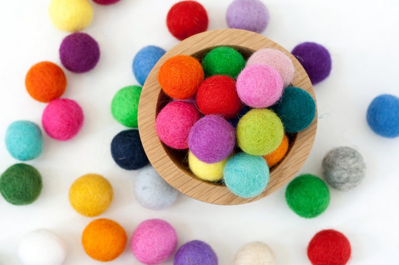 May include: A wooden bowl filled with colorful felt balls. The bowl is surrounded by more felt balls in various colors, including red, blue, green, yellow, orange, pink, purple, and white.