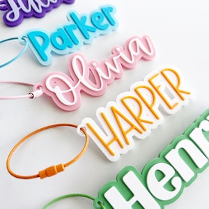 May include: A collection of five personalized luggage tags featuring raised lettering in purple, light blue, pink, orange, and green. Each tag includes a matching colored wire loop fastener. The names displayed are Julia, Parker, Olivia, Harper, and Henry.